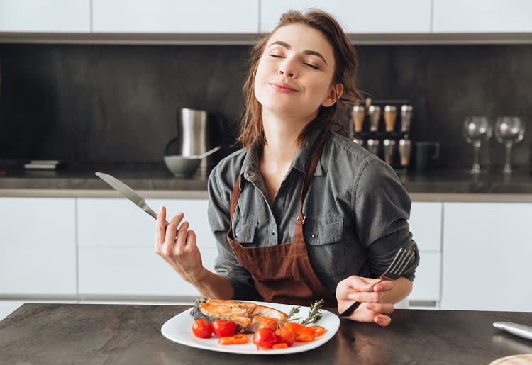 10 Fat-Burning, High-Protein Meals Pescatarians Love Smiling woman holding knife and fork sitting in front of plate of fish and cherry tomatoes